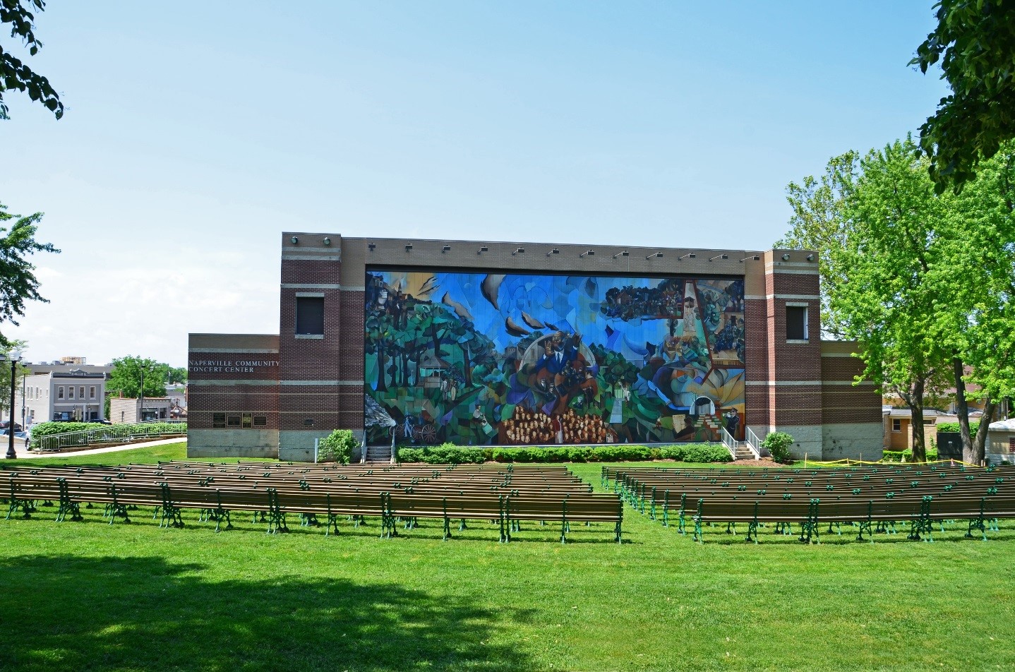 park benches lined up in green grass before a building featuring a mural and a stage
