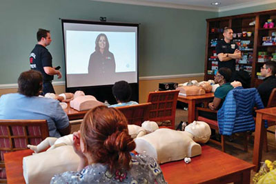 citizens seated at tables watch a CPR video as firefighter/paramedics stand at the front of the room 