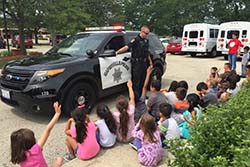 An officer stands outside a squad car while children sit on a curb in front of him, some with hands raised as if asking questions