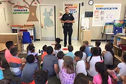 A uniformed officer stands before children seated on the floor in a classroom
