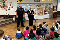 A crime prevention specialist and a uniformed officer stand in front of children seated on the floor of a classroom
