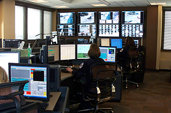 a woman sits at a terminal surrounded by computer monitors, phones and television monitors