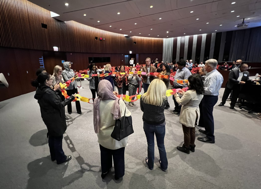 Community members stand in a circle in council chambers holding a paper chain symbolizing inclusion and connection
